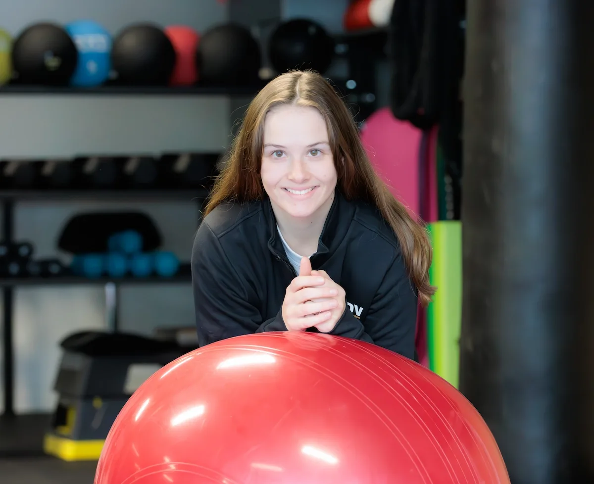 Sofie Crombez leunt lachend op een rode fitnessbal in de trainingsruimte van MOOOV
